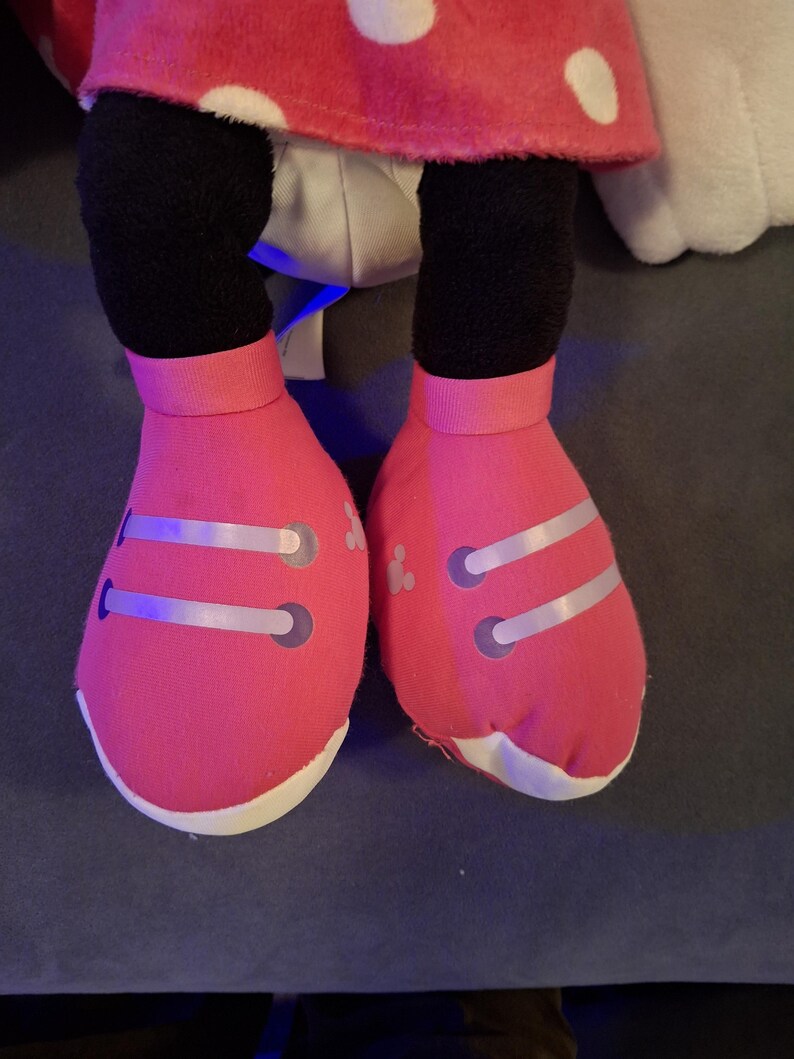 May include: Close-up of a plush toy's legs and feet. The feet are bright pink with white soles and silver accents. The legs are black, and the toy is wearing a red polka-dot skirt.