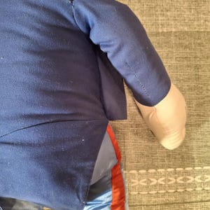 May include: A plush doll wearing a navy blue jacket and light blue pants with a red stripe down the side. The doll's arm is visible, with a tan fabric hand.