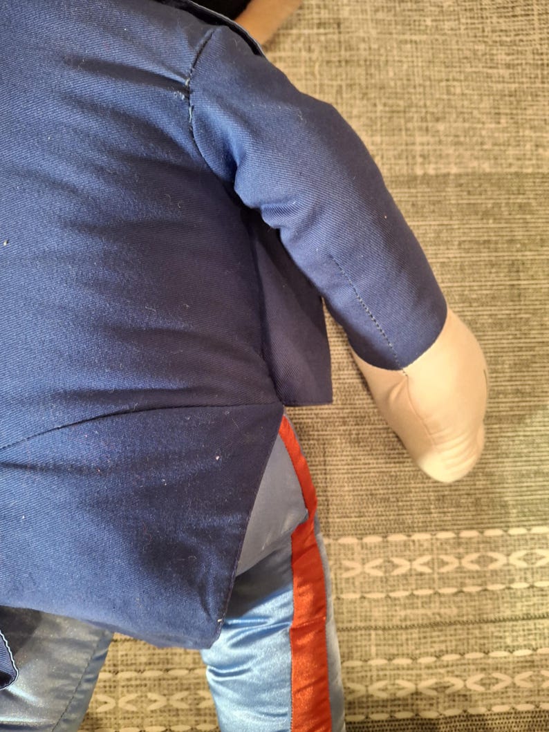 May include: A plush doll wearing a navy blue jacket and light blue pants with a red stripe down the side. The doll's arm is visible, with a tan fabric hand.