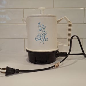 May include: A vintage white electric coffee pot with a black base and handle. The pot features a blue floral design and a black power cord with a two-prong plug. The handle is white and has a ribbed design.