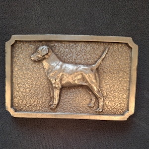 May include: A rectangular metal belt buckle with a raised relief of a dog. The dog is standing, facing left, with its tail up. The buckle has a textured background and a decorative border. The metal has a silver color.