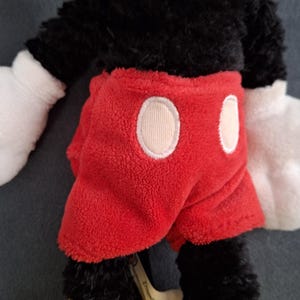 May include: A plush Mickey Mouse toy. The toy has a black body, white gloves, and yellow shoes. It is wearing red shorts with two white circles. The toy is made of soft, fuzzy material.