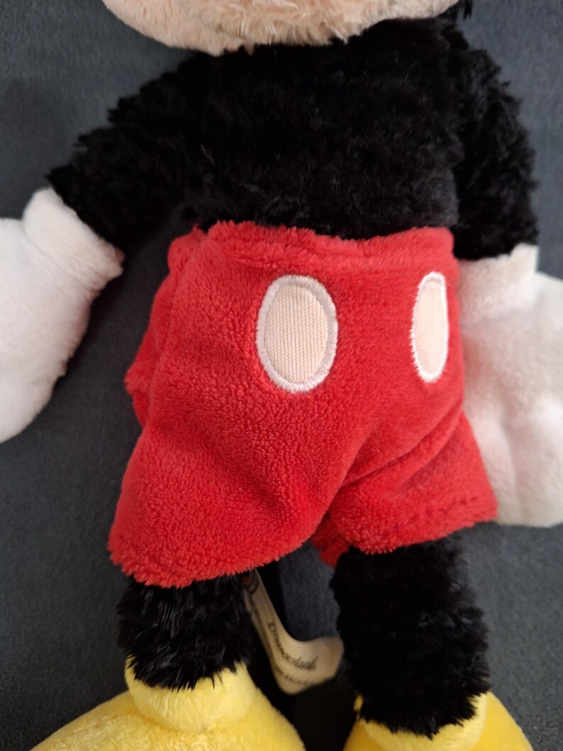 May include: A plush Mickey Mouse toy. The toy has a black body, white gloves, and yellow shoes. It is wearing red shorts with two white circles. The toy is made of soft, fuzzy material.