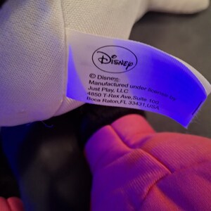 May include: A close-up of a Disney product tag. The tag is white with the Disney logo and text that reads "Disney Manufactured under license by Just Play, LLC". A pink object is in the foreground.
