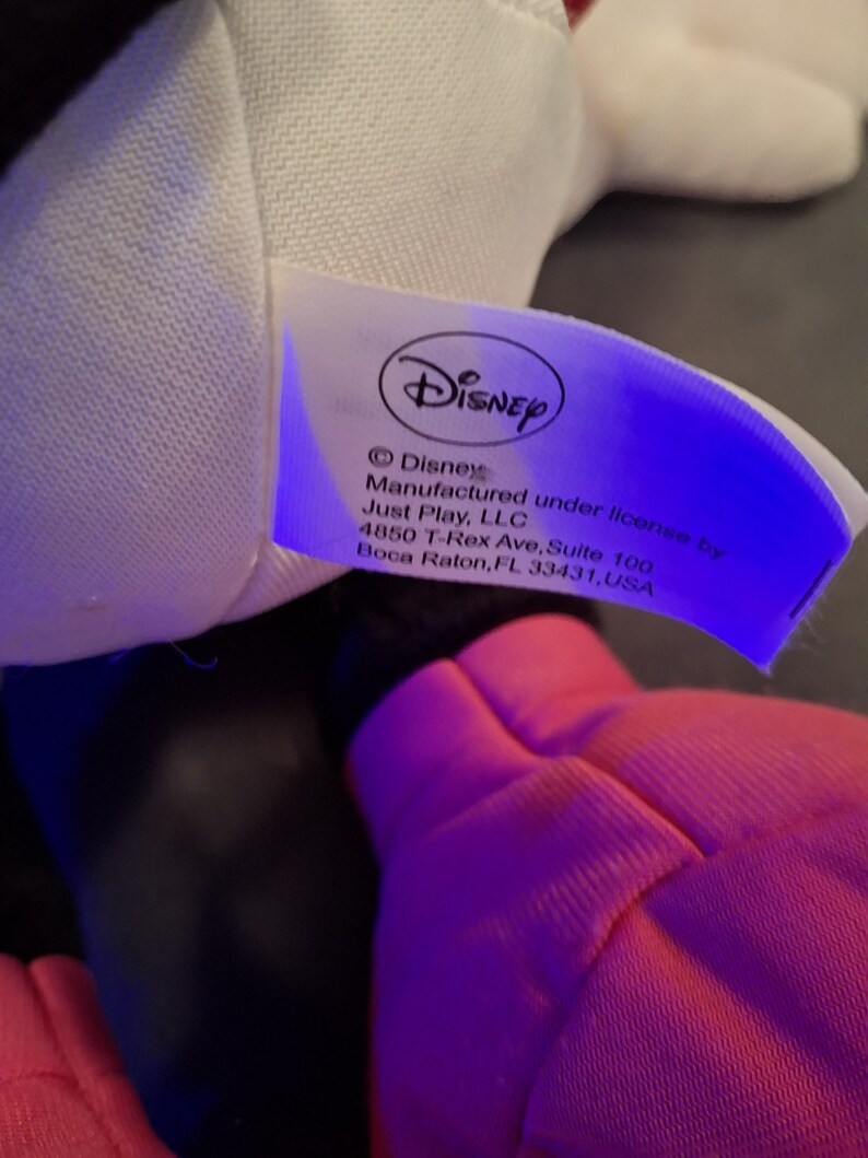May include: A close-up of a Disney product tag. The tag is white with the Disney logo and text that reads "Disney Manufactured under license by Just Play, LLC". A pink object is in the foreground.