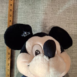 May include: A plush Mickey Mouse doll with black ears, a black nose, and a red bow tie. The doll is wearing a blue jacket. A wooden ruler is next to the doll, measuring approximately 20 inches.