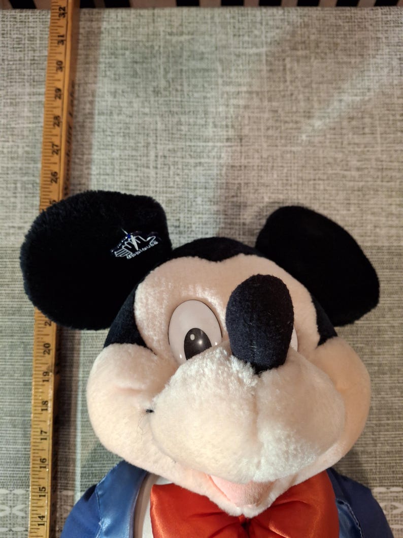May include: A plush Mickey Mouse doll with black ears, a black nose, and a red bow tie. The doll is wearing a blue jacket. A wooden ruler is next to the doll, measuring approximately 20 inches.