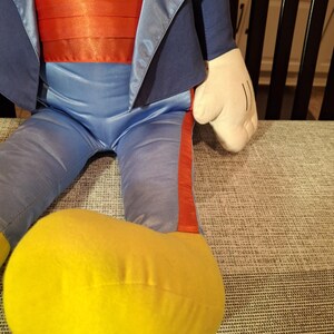 May include: A plush doll featuring a blue suit jacket, pants, and a red cummerbund. The doll has a white torso and hands, with a large yellow foot. The doll is sitting on a gray patterned surface.