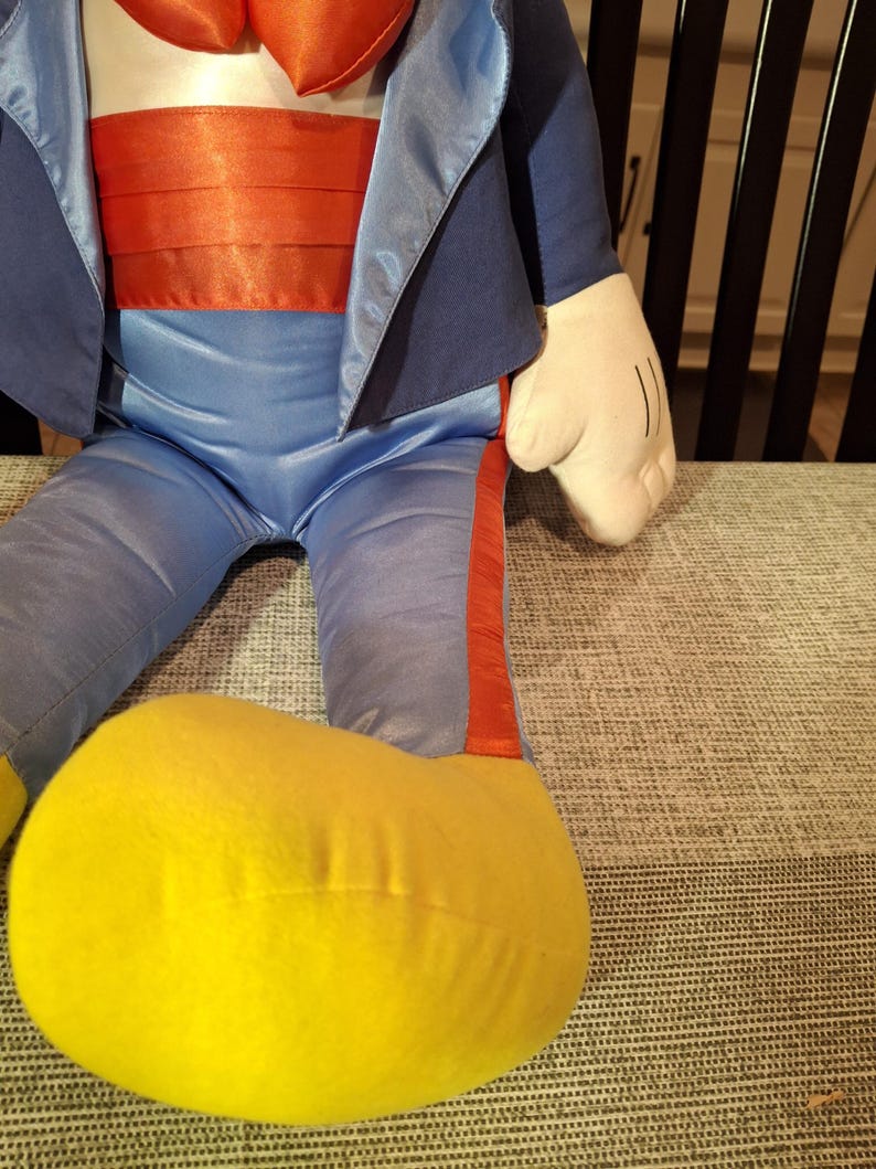 May include: A plush doll featuring a blue suit jacket, pants, and a red cummerbund. The doll has a white torso and hands, with a large yellow foot. The doll is sitting on a gray patterned surface.