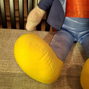 May include: A plush doll with a blue jacket, red sash, and blue pants. The doll has a yellow foot and a cream-colored arm. The doll is sitting on a gray patterned surface.