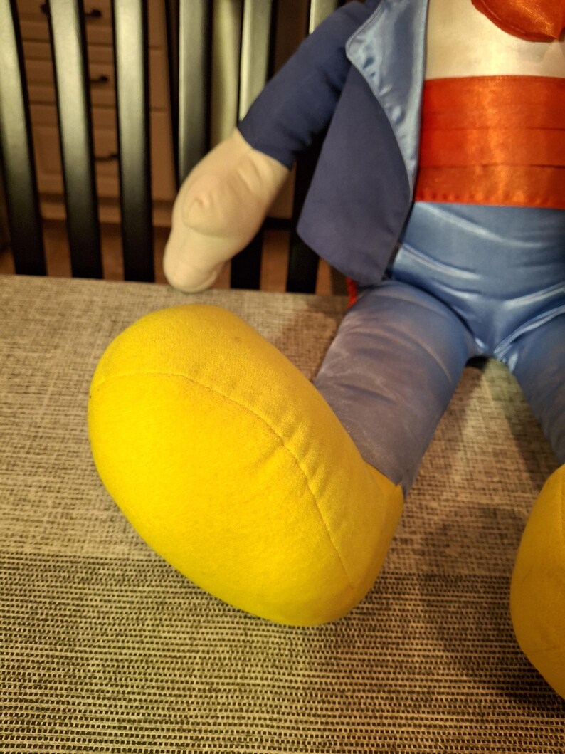 May include: A plush doll with a blue jacket, red sash, and blue pants. The doll has a yellow foot and a cream-colored arm. The doll is sitting on a gray patterned surface.