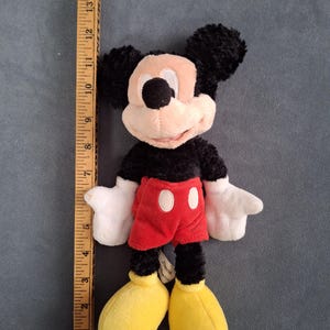 May include: A plush Mickey Mouse toy, approximately 10 inches tall, with black fur, a pink face, and large yellow shoes. He wears red shorts with white buttons and white gloves. The toy is on a gray background.