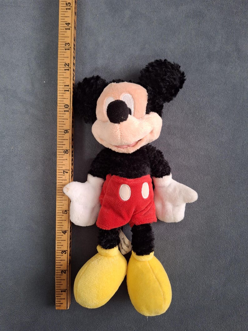 May include: A plush Mickey Mouse toy, approximately 10 inches tall, with black fur, a pink face, and large yellow shoes. He wears red shorts with white buttons and white gloves. The toy is on a gray background.
