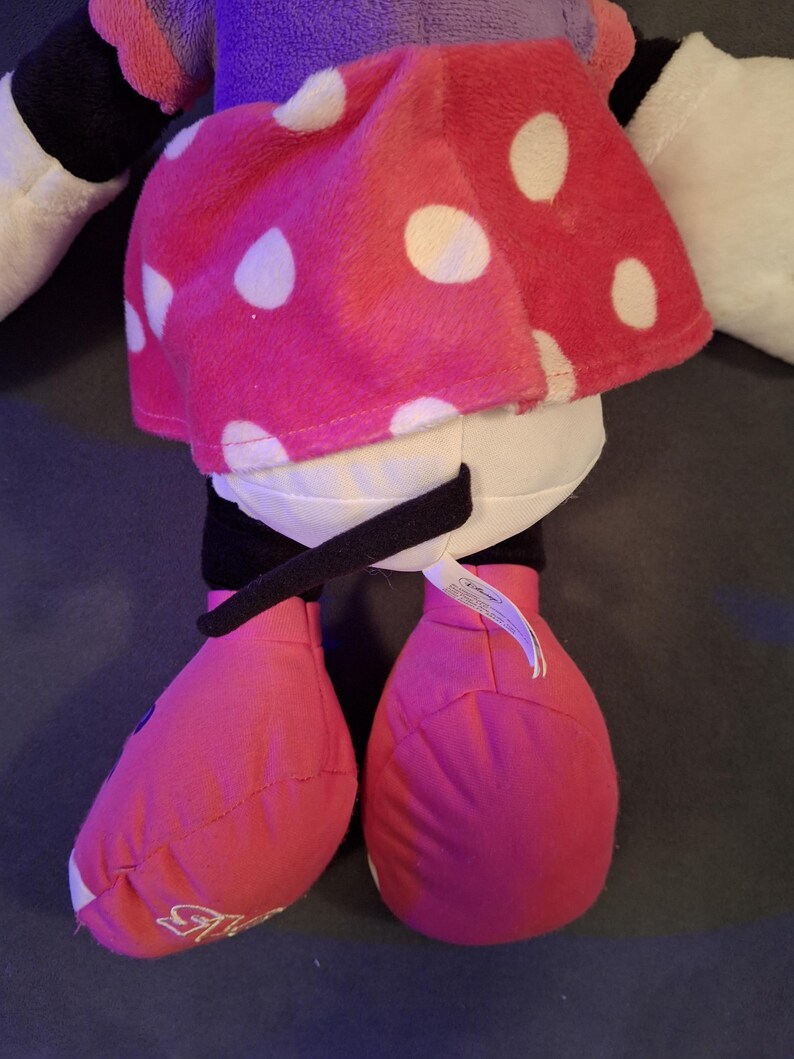 May include: A plush doll featuring a pink skirt with white polka dots, pink shoes, and black accents. The doll has white arms and a purple top. A black belt-like detail is visible. The doll is a popular character.