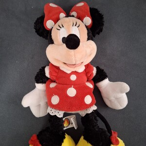 May include: A plush Minnie Mouse doll. The doll has a red dress with white polka dots, a matching bow, and yellow shoes. The doll has black fur, white gloves, and a smiling face. A tag is attached to the doll.
