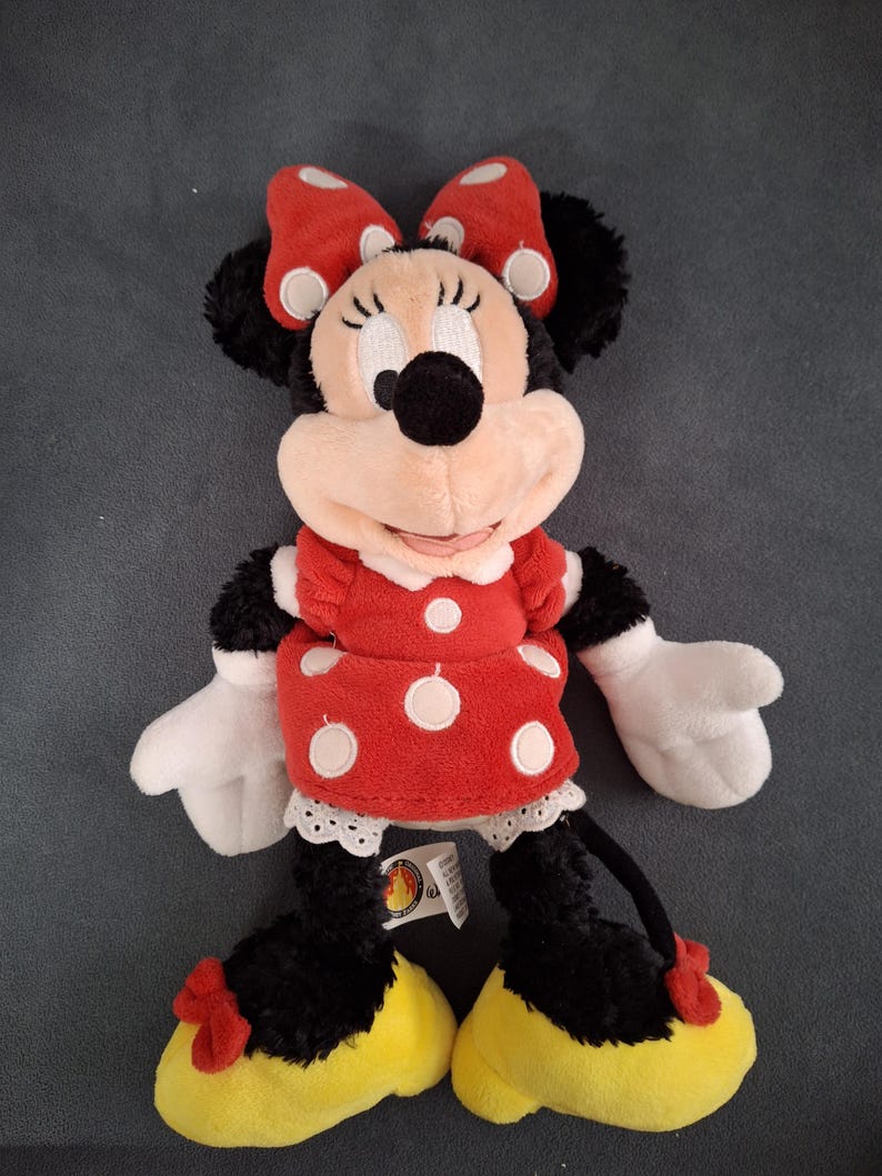 May include: A plush Minnie Mouse doll. The doll has a red dress with white polka dots, a matching bow, and yellow shoes. The doll has black fur, white gloves, and a smiling face. A tag is attached to the doll.