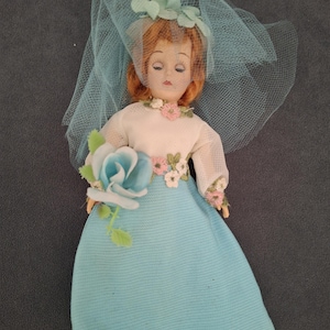 May include: A vintage doll dressed as a bride, featuring a light blue dress and veil. The doll has red hair and is adorned with floral accents in pink, white, and green. It holds a blue rose bouquet, evoking a classic wedding theme.