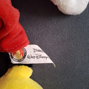 May include: Close-up of a plush toy featuring red, yellow, and white elements. The toy has a Disney Parks tag attached. The tag includes the text "Disney" and "Walt Disney".