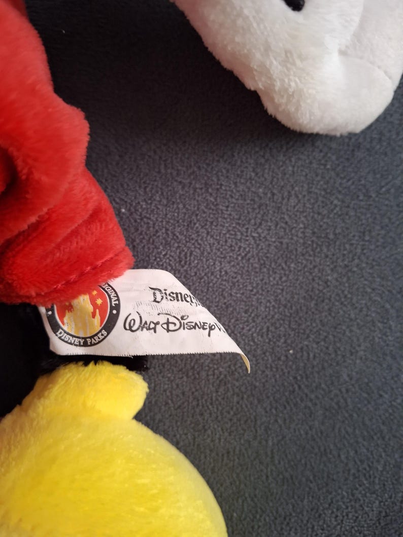May include: Close-up of a plush toy featuring red, yellow, and white elements. The toy has a Disney Parks tag attached. The tag includes the text "Disney" and "Walt Disney".