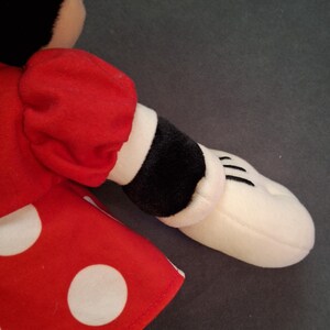 May include: A soft toy with a red dress and white polka dots, a black sleeve with a white cuff, and a white hand with black lines. The toy has a black head and is likely a cartoon character.