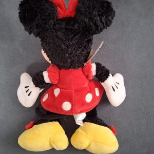 May include: A plush Minnie Mouse doll, viewed from the back, features black fur, a red polka-dot dress, and a red bow. The doll has white gloves and yellow shoes. A tag is attached.