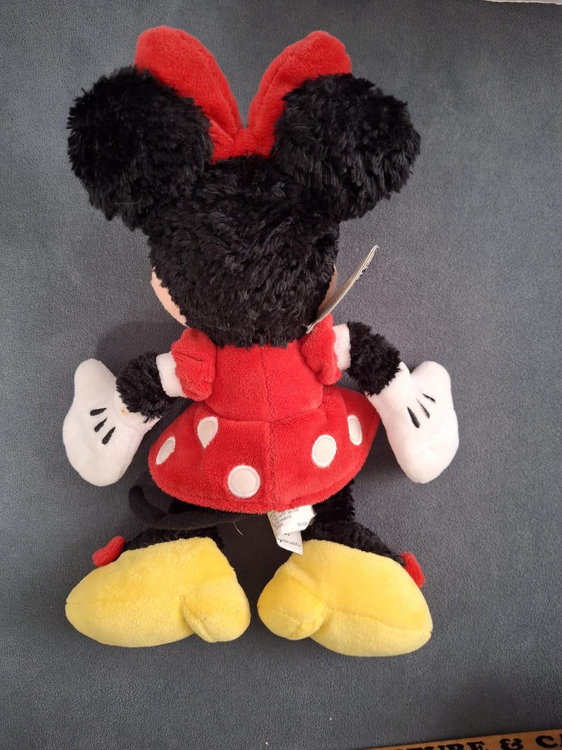 May include: A plush Minnie Mouse doll, viewed from the back, features black fur, a red polka-dot dress, and a red bow. The doll has white gloves and yellow shoes. A tag is attached.