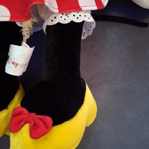 May include: Close-up of a plush toy featuring a red and white polka dot skirt with white lace trim. The toy has black legs, yellow shoes with red bows, and a Disney Store tag.