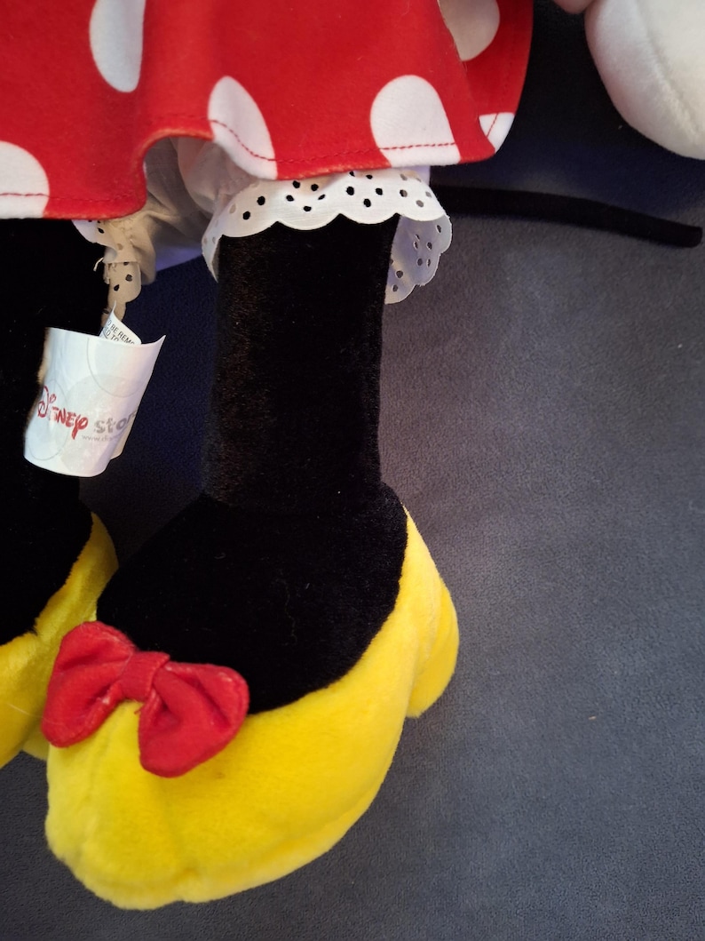 May include: Close-up of a plush toy featuring a red and white polka dot skirt with white lace trim. The toy has black legs, yellow shoes with red bows, and a Disney Store tag.