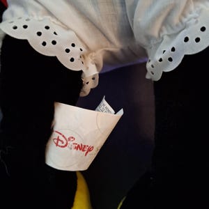 May include: Close-up of a plush toy with black legs and a yellow lower body. The toy has a white ruffled garment with eyelet detailing. A Disney tag is visible.