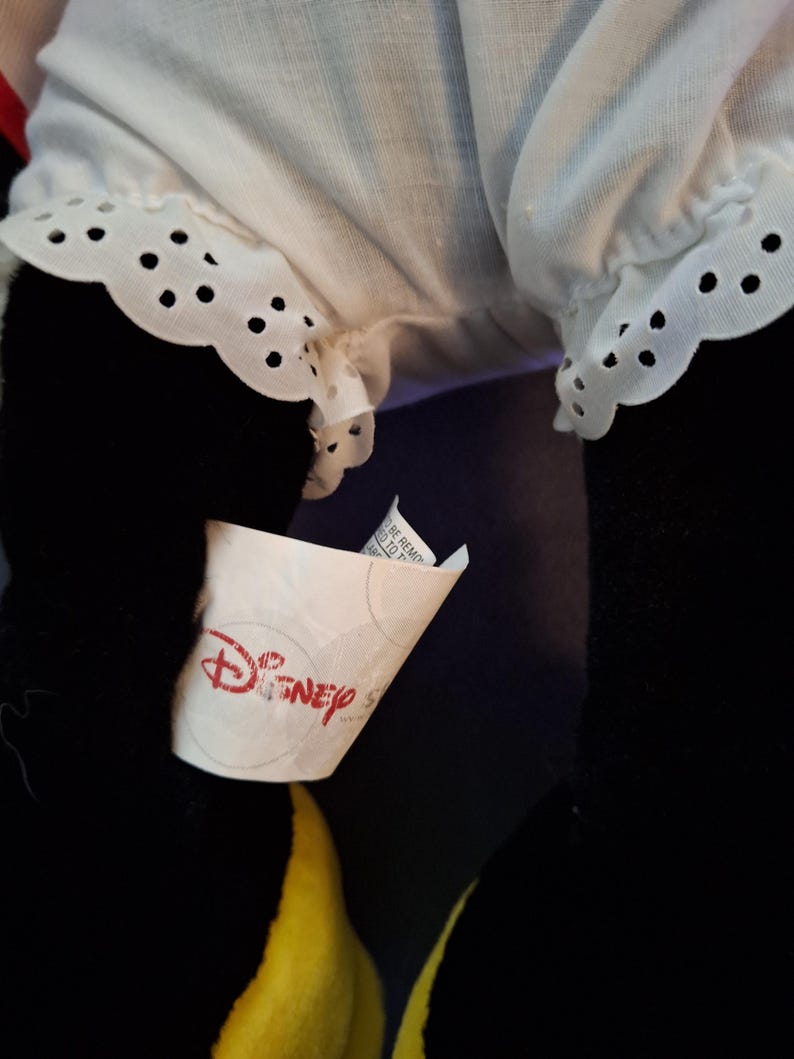 May include: Close-up of a plush toy with black legs and a yellow lower body. The toy has a white ruffled garment with eyelet detailing. A Disney tag is visible.