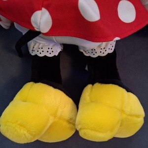 May include: A soft toy featuring a red dress with white polka dots, white ruffled bloomers, black legs, and large yellow feet. The toy's design is reminiscent of a popular cartoon character.