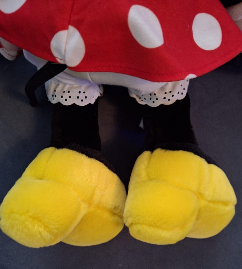 May include: A soft toy featuring a red dress with white polka dots, white ruffled bloomers, black legs, and large yellow feet. The toy's design is reminiscent of a popular cartoon character.