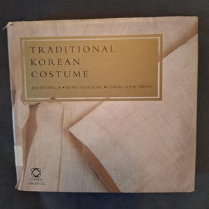 May include: A square hardcover book titled Traditional Korean Costume by Lee Kyung Ja, Hong Na Young, and Chang Sook Hwan. The cover features a light beige fabric texture with a gold rectangular panel containing the title and author names.