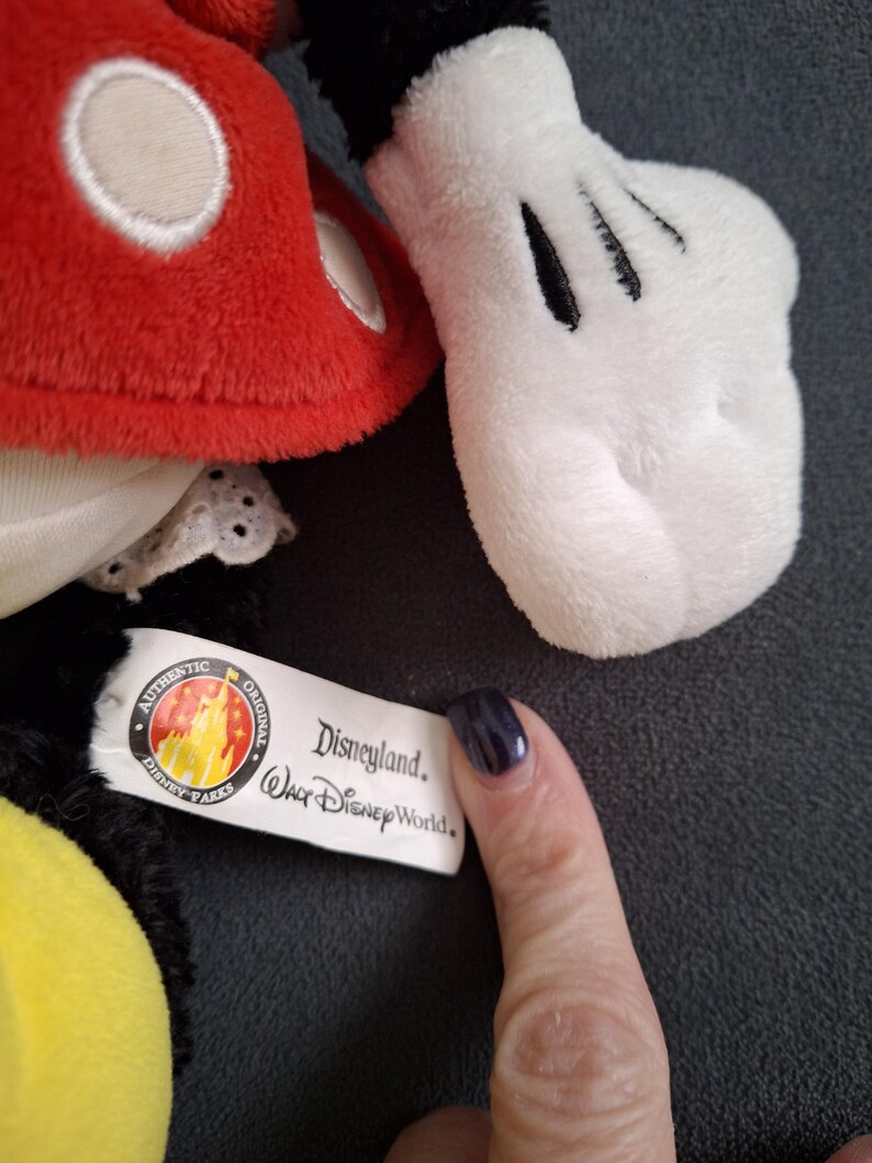 May include: Close-up of a Mickey Mouse plush toy. The toy features a red head with white accents, a white glove with black details, and a tag that reads "Disneyland Walt Disney World."