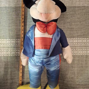 May include: A plush Mickey Mouse doll wearing a blue tuxedo jacket, pants, and an orange bow tie. The doll has black ears, a cream-colored face, and yellow shoes. The doll is approximately 24 inches tall.