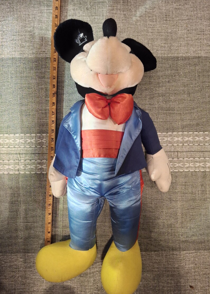 May include: A plush Mickey Mouse doll wearing a blue tuxedo jacket, pants, and an orange bow tie. The doll has black ears, a cream-colored face, and yellow shoes. The doll is approximately 24 inches tall.