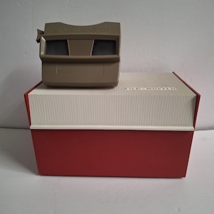 May include: A taupe View-Master stereoscope viewer resting on a rectangular storage case. The case features a red base and a white, vertically ribbed lid with the word View-Master embossed on the top surface. The viewer has two lenses.