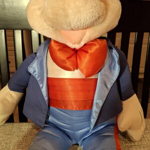 May include: A plush toy featuring a cartoon character with a large, round head, black ears, and a white face. The character is dressed in a blue suit with a satin lapel, a red bow tie, and a red cummerbund. The toy is seated.