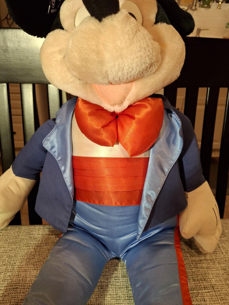 May include: A plush toy featuring a cartoon character with a large, round head, black ears, and a white face. The character is dressed in a blue suit with a satin lapel, a red bow tie, and a red cummerbund. The toy is seated.