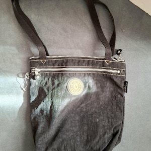 May include: Black nylon crossbody bag with a textured appearance. Features a top zipper, a small round logo, and a long black strap. The bag has a small tag on the side and a silver zipper.