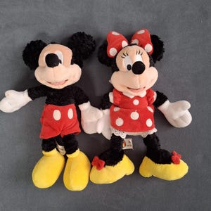 May include: Two plush Disney character dolls, Mickey Mouse and Minnie Mouse, are shown. Mickey wears red shorts with white buttons and yellow shoes. Minnie wears a red polka-dot dress, a red bow, and yellow shoes.