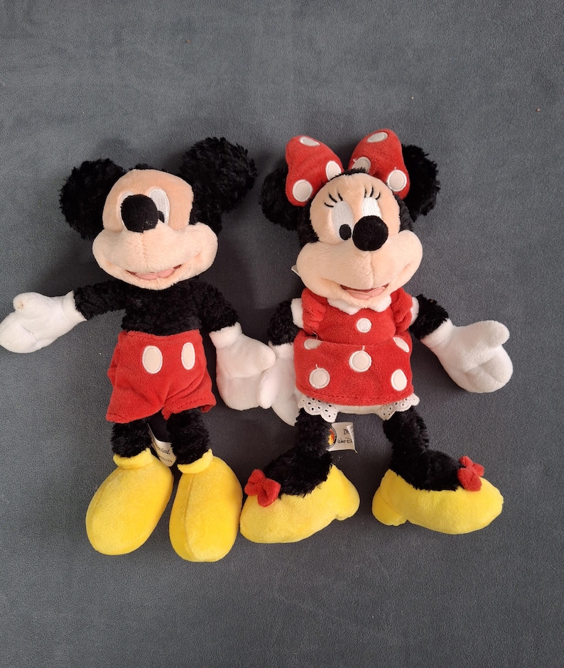 May include: Two plush Disney character dolls, Mickey Mouse and Minnie Mouse, are shown. Mickey wears red shorts with white buttons and yellow shoes. Minnie wears a red polka-dot dress, a red bow, and yellow shoes.