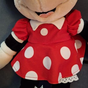 May include: A Minnie Mouse soft toy, dressed in a red dress with white polka dots, a white collar, and a white petticoat. The doll has a beige face, black ears, and black arms with white cuffs. A Disney tag is visible.