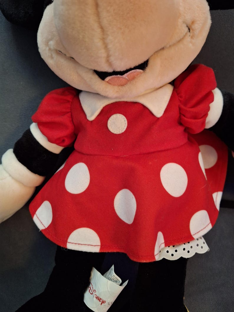 May include: A Minnie Mouse soft toy, dressed in a red dress with white polka dots, a white collar, and a white petticoat. The doll has a beige face, black ears, and black arms with white cuffs. A Disney tag is visible.