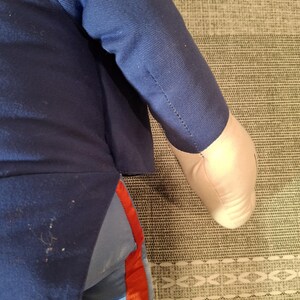 May include: A plush doll wearing a navy blue jacket and light blue pants with a red stripe. The doll's arm is visible, with a white hand. The doll is lying on a patterned surface.