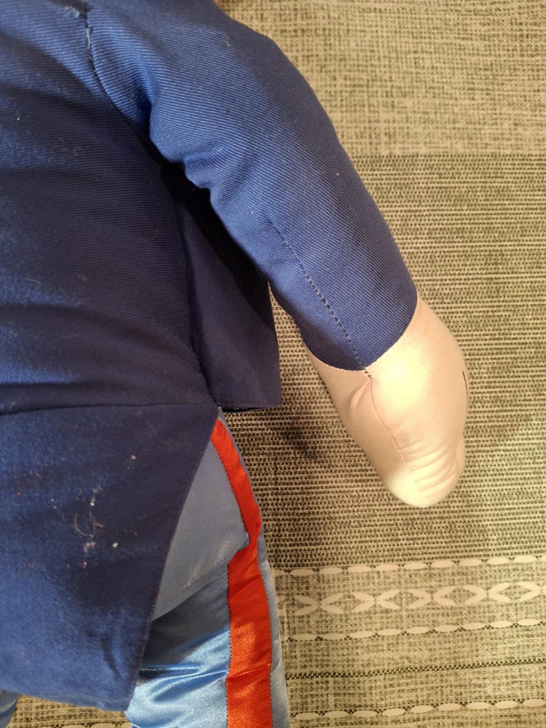 May include: A plush doll wearing a navy blue jacket and light blue pants with a red stripe. The doll's arm is visible, with a white hand. The doll is lying on a patterned surface.