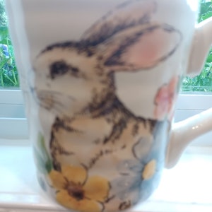 May include: White ceramic mug with a brown rabbit illustration, surrounded by yellow, blue, and pink flowers.