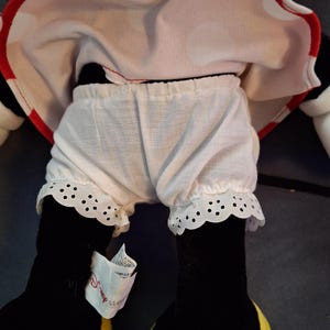May include: A Minnie Mouse soft toy, wearing a white and red polka dot dress with red trim. The doll has white bloomers with a scalloped lace trim. The doll's black legs have yellow shoes. A Disney tag is attached.