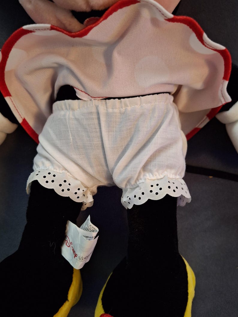 May include: A Minnie Mouse soft toy, wearing a white and red polka dot dress with red trim. The doll has white bloomers with a scalloped lace trim. The doll's black legs have yellow shoes. A Disney tag is attached.