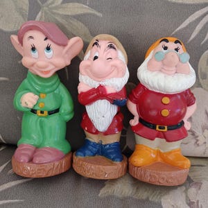 May include: Three plastic figurines of the seven dwarfs from the Disney movie Snow White. The dwarfs are named Dopey, Grumpy, and Doc. The figurines are in a seated position and are wearing their signature clothing.